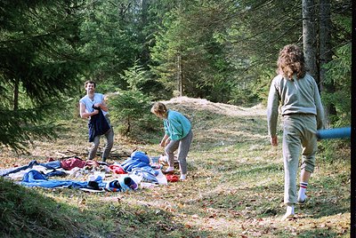 Three individuals in a forested area, likely mid-1970s, organizing camping gear. Lush pine trees frame the scene, with a mix ...