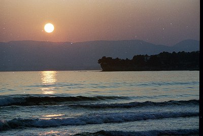 Golden sunset over calm waters with distant island silhouette and misty mountain range. Warm tones reflect on gentle waves, c...