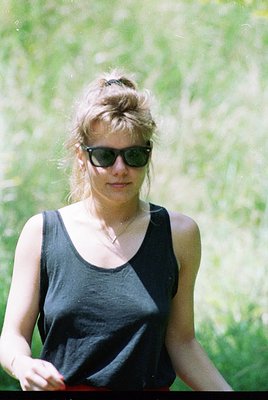 Vintage-style portrait of a woman in a sleeveless black top, sunglasses, and red shorts, set against blurred greenery. Mid-20...