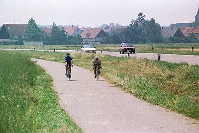 Three cyclists ride along a rural gravel path beside a road with vintage cars (, , ). Traditional Dutch farmhouses with red-t...