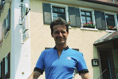 Vintage photo of a man in a blue polo shirt with a logo, standing outside a European-style building with shuttered windows an...