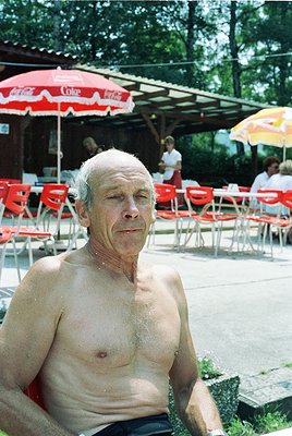 Shirtless man seated outdoors under red Coca-Cola branded umbrellas, surrounded by red plastic chairs. Mid-20th century Europ...