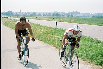 Two cyclists race on a rural road, one shirtless in black shorts, the other in a white/red jersey. Classic road bike frames a...