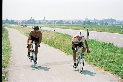 Two cyclists race on a rural road, one shirtless in black, the other in a white/red kit. Classic road bike frames and drop ha...