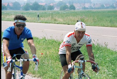 Two cyclists in vintage road gear race on a rural road, 1970s. Rider left wears blue jersey with white stripes, sunglasses, a...