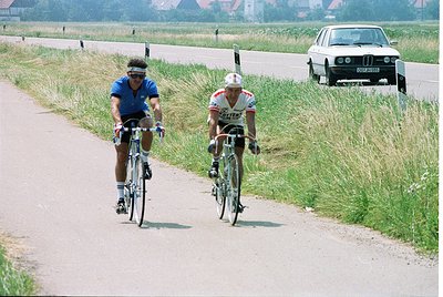 Two cyclists race on a rural road, 1970s-80s era road bikes with drop handlebars. Rider left wears blue kit, right sports whi...