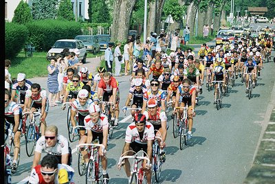 Packed cycling race with riders in vintage jerseys and helmets, likely 1970s–1980s. Spectators line both sides of a tree-line...