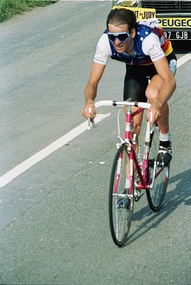 Cyclist in a 1980s road race jersey (Peugeot team) leaning forward on a vintage road bike, gripping handlebars during a sprin...