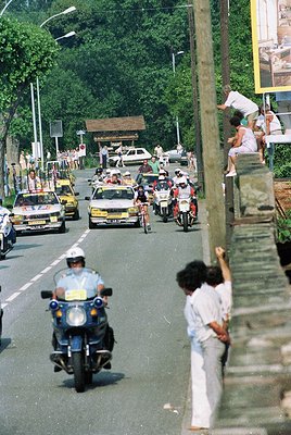 Vintage street scene with motorcade: police motorcycles, vintage cars, and spectators lining a tree-lined road. Mid-20th cent...