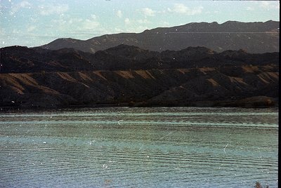 Vintage landscape shot of a serene lake bordered by rugged, forested mountains under a clear sky. The water’s surface reflect...