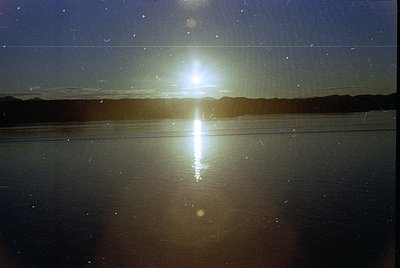 Sunburst reflection on calm water, likely a lake or coastal inlet, with lens flare creating symmetrical light patterns. Horiz...
