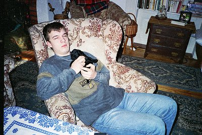 Vintage 1990s indoor portrait of a young man lounging in a floral-patterned armchair, holding a black flip-style phone. Surro...
