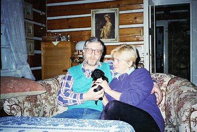 Vintage indoor portrait of two adults holding a black-and-white cat on a floral-patterned sofa in a rustic wooden cabin. Fram...