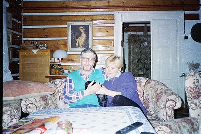 Vintage wooden cabin interior with two adults seated on floral-patterned sofas, examining a small handheld device. Warm light...