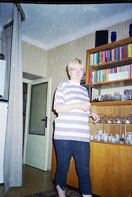Mid-century interior featuring a woman in a striped shirt and dark trousers, standing beside a wooden bookshelf displaying co...