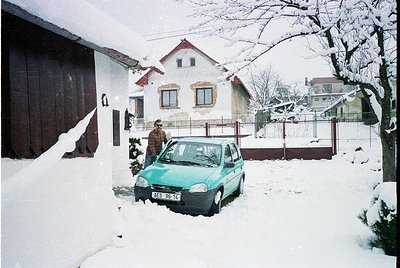 Snow-covered residential street with 1990s-era compact car (turquoise, likely Škoda) parked beside a light-colored house. Man...