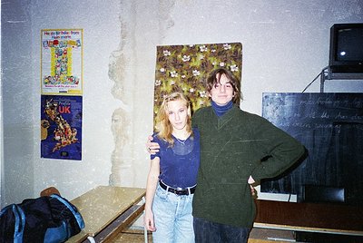 Two individuals pose indoors in a lived-in space, likely a dorm or shared flat, circa 1990s. The woman wears a blue top with ...