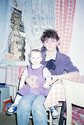 A woman in a striped blouse and jeans holds a toddler in a purple dress, seated on a wooden chair in an indoor setting. Behin...