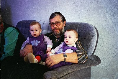 A man in glasses holds two infants in a vintage armchair, likely mid-1980s–1990s. Both children wear layered, patterned cloth...