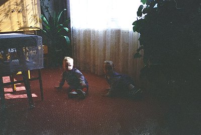 Vintage indoor scene of two young children playing on a red carpeted floor, likely mid-20th century. Child on left sits near ...