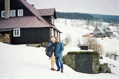 Two individuals pose in winter attire—one in a blue jacket, beige pants, and a backpack; the other in a dark jacket and jeans...
