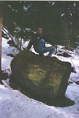 Man poses atop moss-covered boulder in snow-covered forest, wearing dark jacket, jeans, and a logoed shirt. Snow blankets gro...