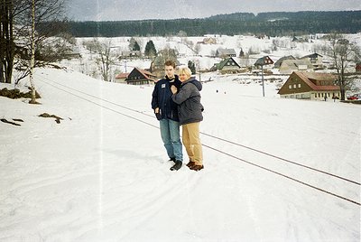 Two individuals embrace on a snow-covered slope beside a railway track, surrounded by a quaint village blanketed in winter. T...