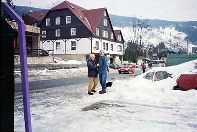 Winter street scene featuring a two-story alpine-style building with white walls, dark trim, and red roof. Two men in winter ...