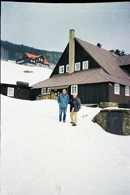 Two men in winter attire pose in front of a dark wooden alpine chalet with a steep gabled roof, surrounded by snow-covered te...