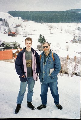 Two men pose in winter attire on a snowy hillside overlooking a quaint village. The younger wears a purple scarf, dark jacket...