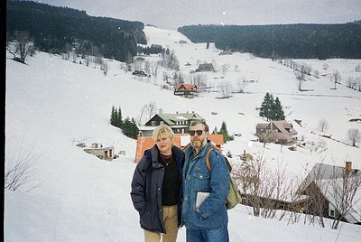 Two individuals pose on a snow-covered slope in a winter village, surrounded by wooden chalets with red roofs. The man wears ...