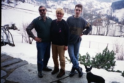 Three individuals pose on a snow-covered terrace in a mountainous region, likely the Alps. The man on the left wears a stripe...