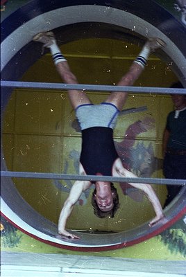 Upside-down gymnastics routine on a horizontal bar, likely in a vintage gymnasium. The athlete wears a striped sleeveless top...