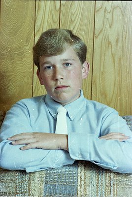 Portrait of a young boy in a light blue dress shirt and white tie, seated against a wooden panelled wall. His expression is n...