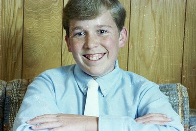 Young boy in a light blue dress shirt and white tie, posing indoors against a wooden panelled wall. Mid-20th century formal p...