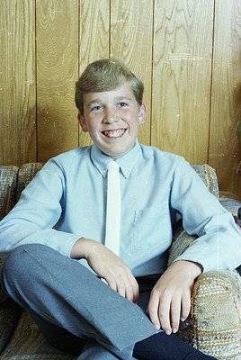 Portrait of a young boy in a light blue dress shirt, dark tie, and trousers, seated on a woven armchair against wooden paneli...