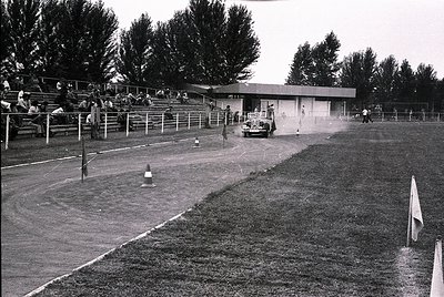 Vintage black-and-white shot of a dirt track racing event, featuring a lone driver in a classic open-wheel car navigating a t...