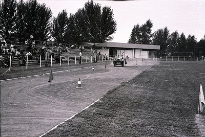 Mid-20th century auto racing track with grandstand seating. A vintage car races on a dirt oval, kicking up dust. Spectators i...