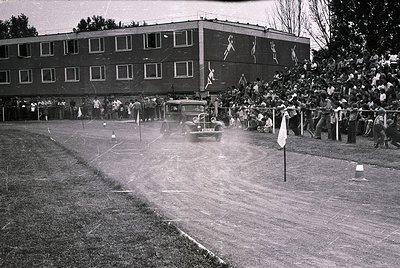 Vintage black-and-white shot of a mid-20th-century auto race on a dirt track. A vintage car speeds past spectators lined behi...