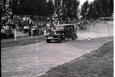 Vintage black-and-white photo of a classic 1930s-era sedan navigating a muddy track during a slalom race. Spectators line woo...