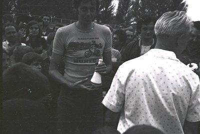 Cyclist in 1970s racing jersey receiving trophy from official at outdoor event. Crowd in background, suggesting post-race cel...