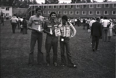 Three men pose outdoors in 1970s-era attire—striped polo shirts, rolled-up sleeves, and name tags—holding what appear to be e...