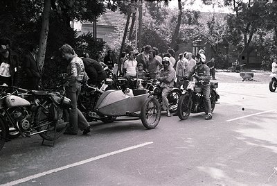 Vintage sidecar motorcycles lined up on a quiet road, surrounded by mid-century attire. Riders in helmets and leather jackets...