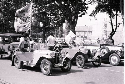 Vintage 1930s road rally with classic cars and vintage flags. Participants wave banners, one featuring a bicycle logo, in a l...