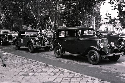 Vintage street scene featuring three classic cars (1930s-1940s) parked on cobblestone pavement. Open-top convertible on left,...