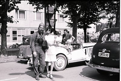 Vintage black-and-white street scene featuring a man in military uniform (likely 1940s–1950s) posing with a woman in a floral...
