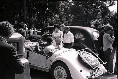 Vintage mid-century roadside gathering featuring classic convertibles and casual 1950s-60s attire. Group of people in light s...