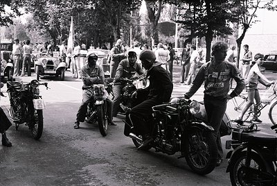 Classic black-and-white street scene featuring vintage motorcyclists in protective gear, likely 1960s–1970s. Riders pose on p...