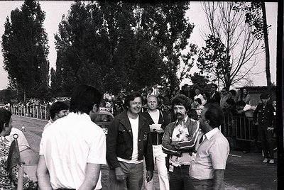 Black-and-white street scene featuring a group of five adults in casual 1970s attire—leather jackets, striped shirts, and bel...
