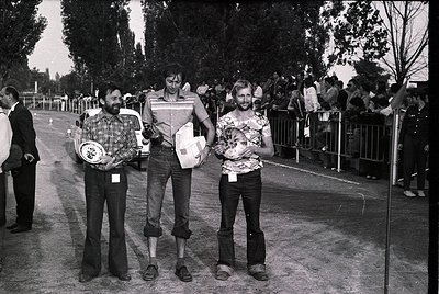 Three men pose holding framed plaques at an outdoor event, likely a sports or cultural competition. The central figure wears ...